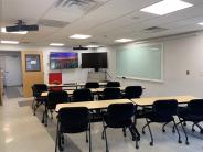Large room with three rectangular tables and black chairs set up classroom style with two large monitors on wall in front
