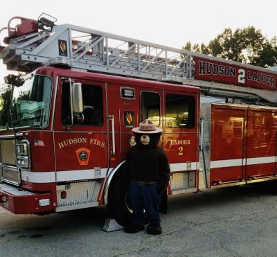Photo of Ladder 2 - red fire truck with ladder across truck.  Smokey the Bear standing next to the drivers door.